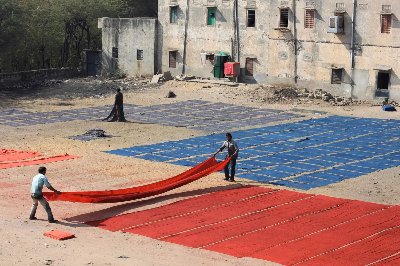 Long lengths of red, blue and purple printed cloth spread across open ground beside village buildings.