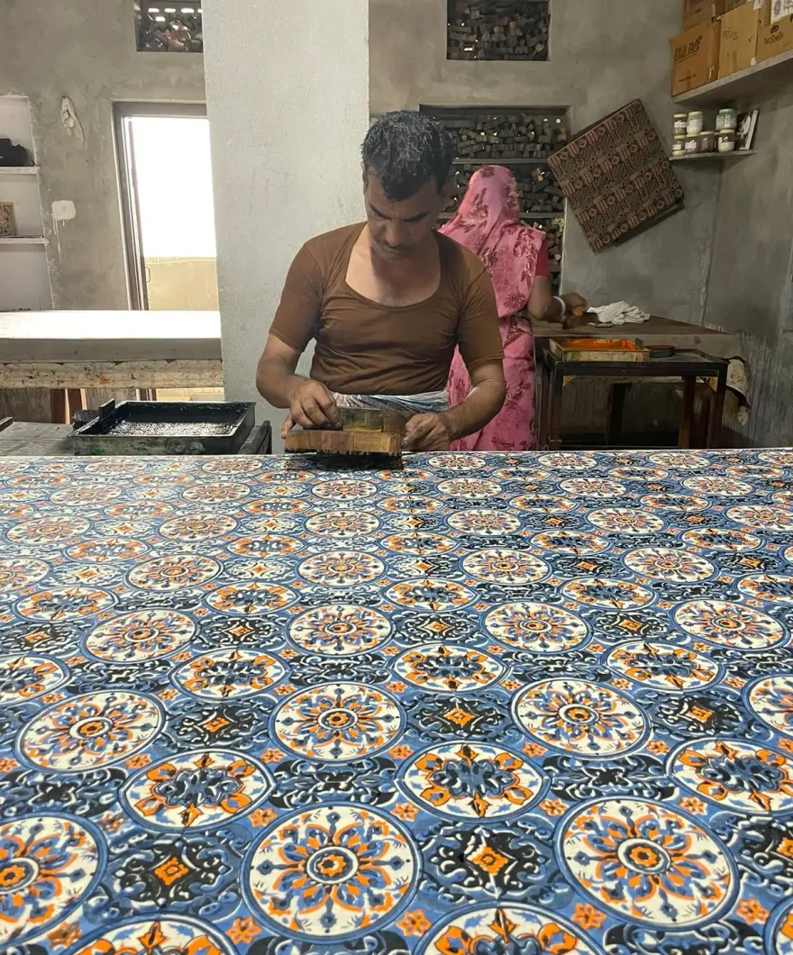 A printer at a long padded table pressing a wooden block onto blue and orange patterned cloth, a tray of colour beside him.