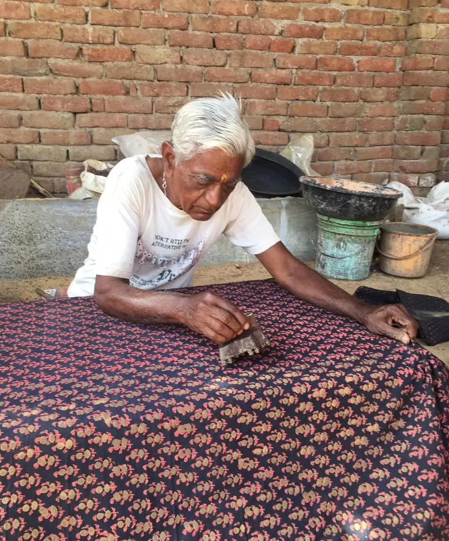 An elderly printer bends over a long table, pressing a small block onto cloth covered in a red and yellow repeat.