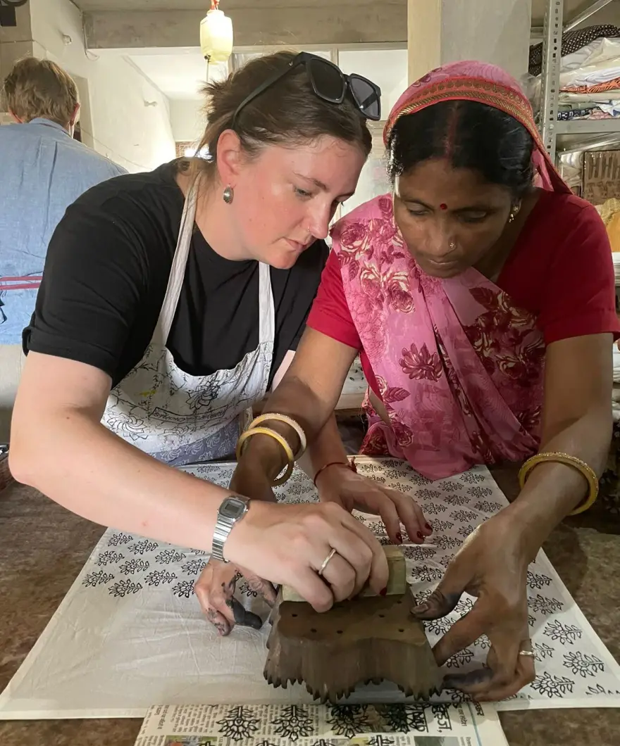Two people at a printing table lining up a carved block on pale cloth, the printed border already run along one edge.