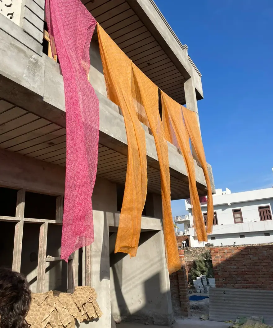 Pink and orange printed scarves hanging from a concrete balcony against a blue sky.