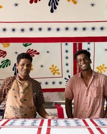 Vijendra and Yash Chhipa at a printing table, with printed cloth hanging behind them.